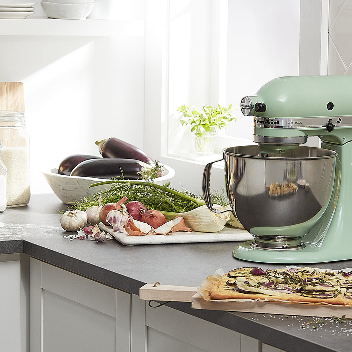 KitchenAid Artisan Series 5-Quart Tilt-Head Stand Mixer in pistachio, on a kitchen counter with fresh produce and a cutting board