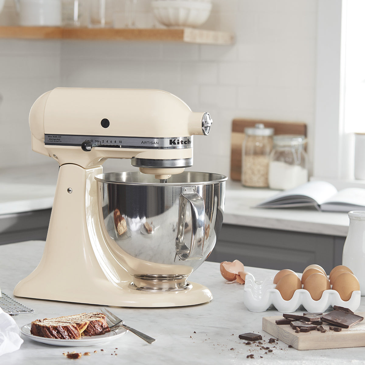 KitchenAid Artisan Series 5-Quart Tilt-Head Stand Mixer in almond cream, on a marble kitchen counter with baking ingredients