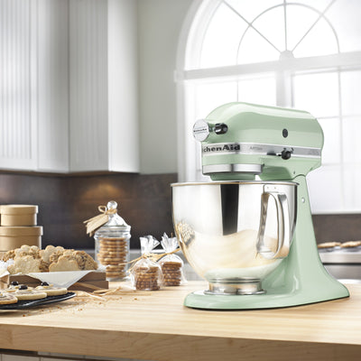 KitchenAid Artisan Series 5-Quart Tilt-Head Stand Mixer in pistachio, on a wooden kitchen counter by a window with baked goods